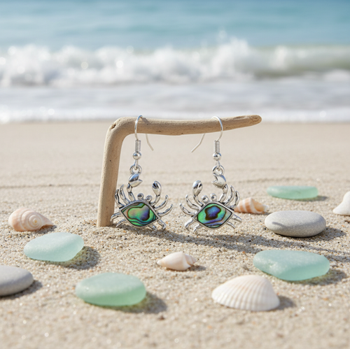 Silver-toned crab dangle earrings with abalone inlay displayed on driftwood stand with sea glass and shells on beach sand - coastal jewelry - Crabby Mermaid