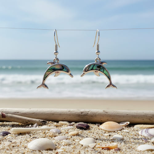 Silver-toned abalone inlay dolphin dangle earrings hanging over driftwood with seashells on beach sand and ocean in background — coastal jewelry — Crabby Mermaid