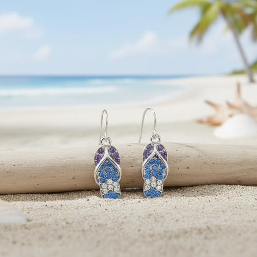 Silver-toned blue crystal flip flop dangle earrings leaning against driftwood on beach sand with ocean and palm trees in background — coastal jewelry — Crabby Mermaid