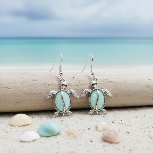 Silver-toned luminous opal sea turtle dangle earrings displayed on driftwood with pastel seashells and dreamy aqua ocean background — coastal jewelry — Crabby Mermaid