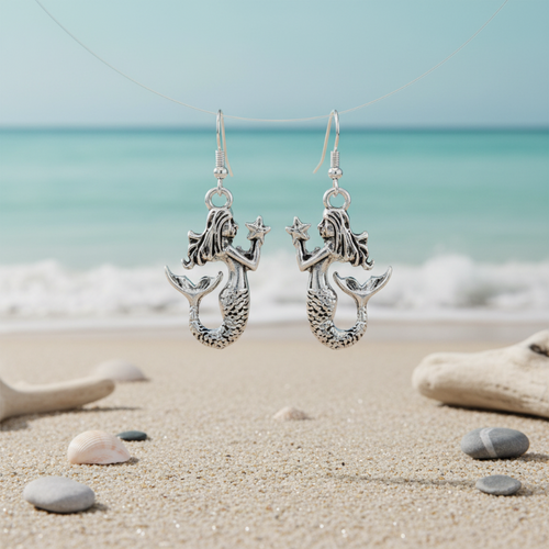 Silver-toned mermaid with starfish dangle earrings floating over beach sand with shells and sea stones and ocean waves in background — coastal jewelry — Crabby Mermaid