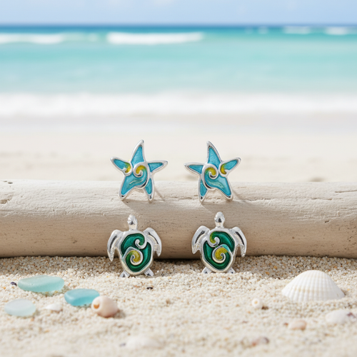Aqua enamel starfish studs and teal enamel sea turtle studs displayed on driftwood with sea glass and shells on beach sand and ocean background - coastal earring set - Crabby Mermaid