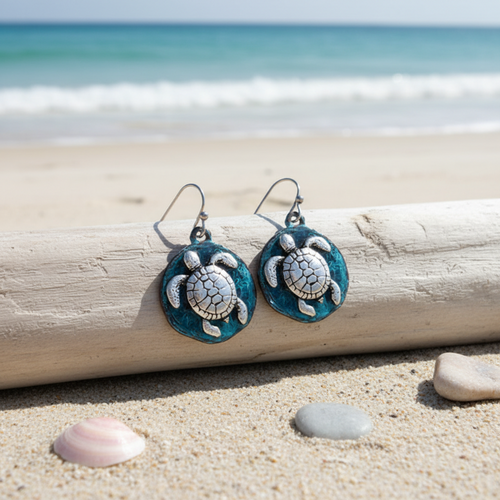 Teal patina sea turtle disc dangle earrings leaning against driftwood with pebbles and shells on beach sand and ocean in background — coastal jewelry — Crabby Mermaid