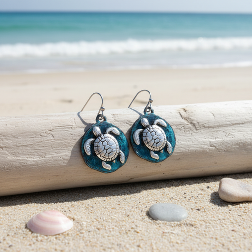 Teal patina sea turtle disc dangle earrings leaning against driftwood with pebbles and shells on beach sand and ocean in background - coastal jewelry - Crabby Mermaid