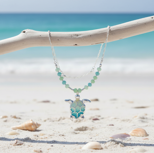 Silver-toned ombre sea turtle beaded necklace hanging from driftwood with shells on beach sand and turquoise ocean in background - coastal jewelry - Crabby Mermaid