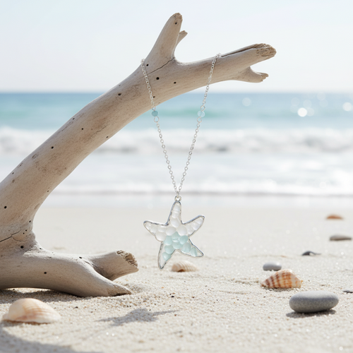 Sea glass starfish pendant necklace hanging from driftwood with ocean waves in background — coastal jewelry — Crabby Mermaid