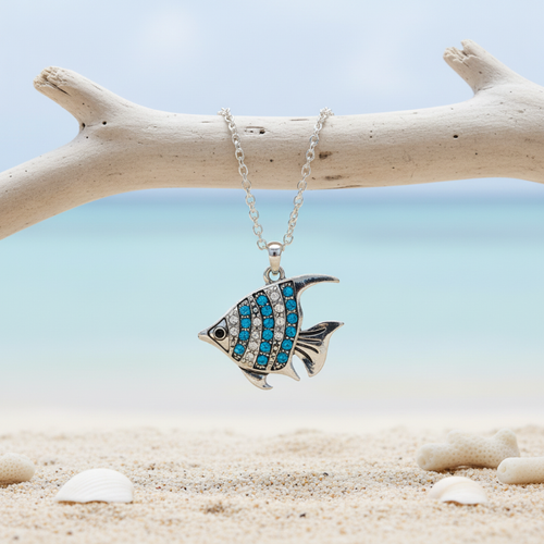 Aqua crystal angel fish necklace hanging from driftwood with shells on beach sand and ocean in background - coastal jewelry - Crabby Mermaid