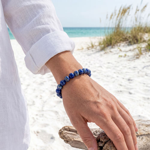 Lapis Lazuli gemstone beaded stretch bracelet on wrist worn with white linen on Florida Gulf Coast beach ‚Äî Crabby Mermaid