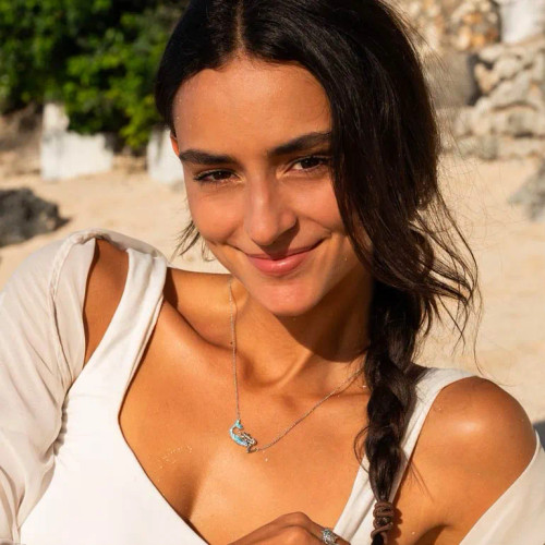 Blue mermaid necklace with Swarovski crystals draped over a seashell on sandy beach, evoking coastal fantasy and ocean-inspired jewelry