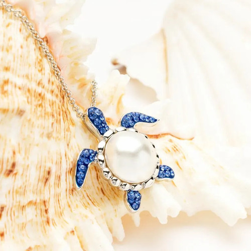 Sterling Silver Pearl Sea Turtle Necklace with Sapphire Crystals - A breathtaking coastal jewelry piece featuring a sea turtle pendant with a lustrous freshwater pearl body, adorned with sapphire blue crystals. The slim sterling silver chain adds elegance, making it a winsome addition from Ocean’s 'Drops of Joy' range. Perfect for embracing the serenity of the deep seas. Made in Ireland
