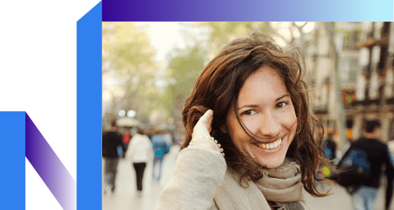 A smiling woman with wavy brown hair stands on a busy street, wearing a scarf.