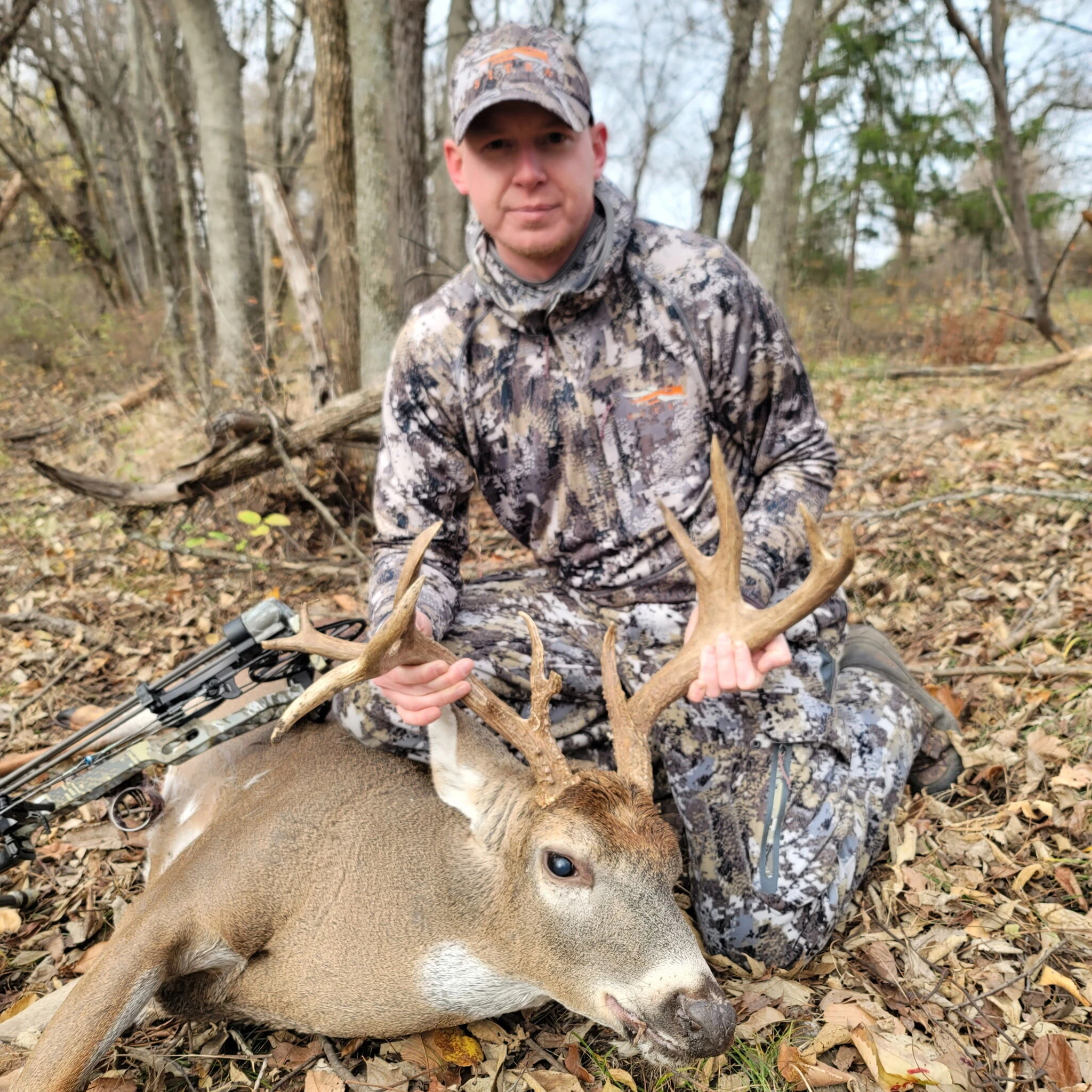 A bowhunter in camouflage kneeling behind a whitetail buck in the woods, with his compound bow on the leaves beside him
