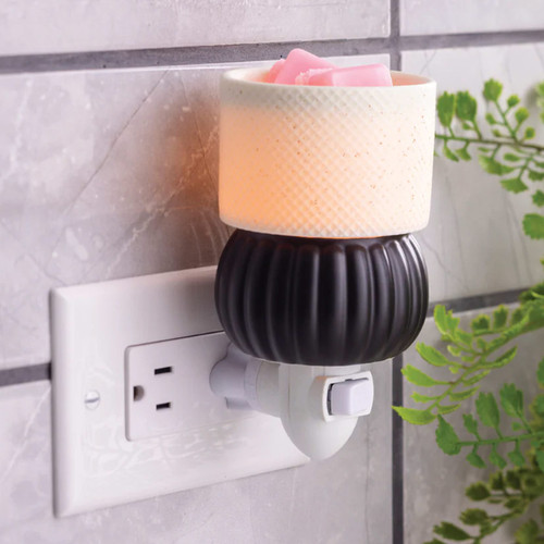 The Luxe Lamp Pluggable Warmer plugged into a kitchen wall outlet, emitting a soft amber glow through the textured ceramic shade while warming pink wax melts