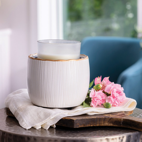 A Golden Ridges Crock Candle Warmer featuring ivory stoneware and a gold rim, warming a jar candle on a decorative tray in a bright, modern living room.