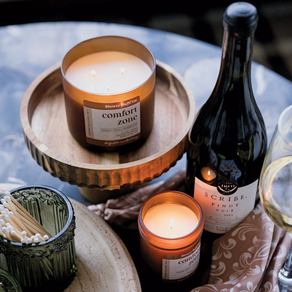 A cozy evening lifestyle scene featuring Slower Days Co. Comfort Zone candles on a wooden pedestal tray alongside a bottle of Pinot Noir and a green glass match holder.