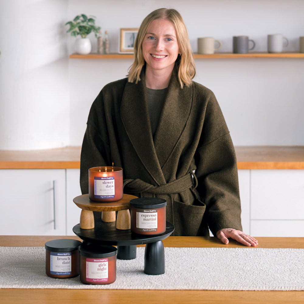 The founder of Slower Days Co. posing with a signature collection of amber glass candles, arranged on decorative tiered stands in a cozy home setting.