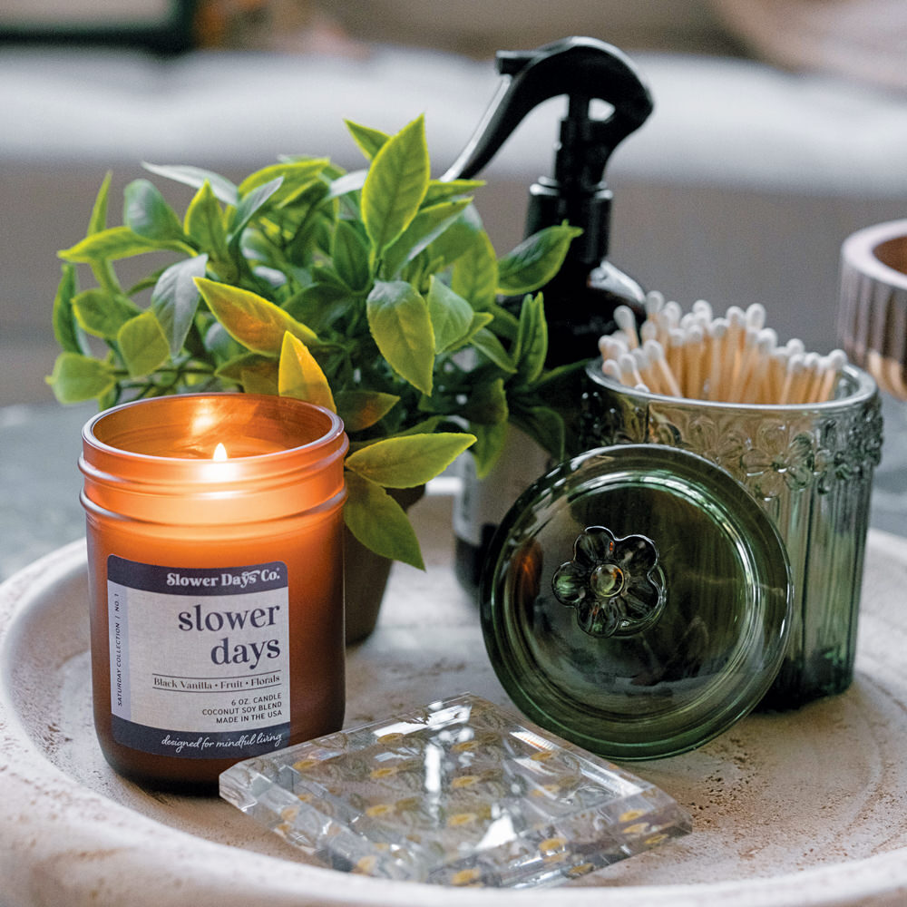 A cozy home decor arrangement featuring a lit Slower Days amber glass candle, a green potted plant, and decorative glass accessories on a textured stone tray.