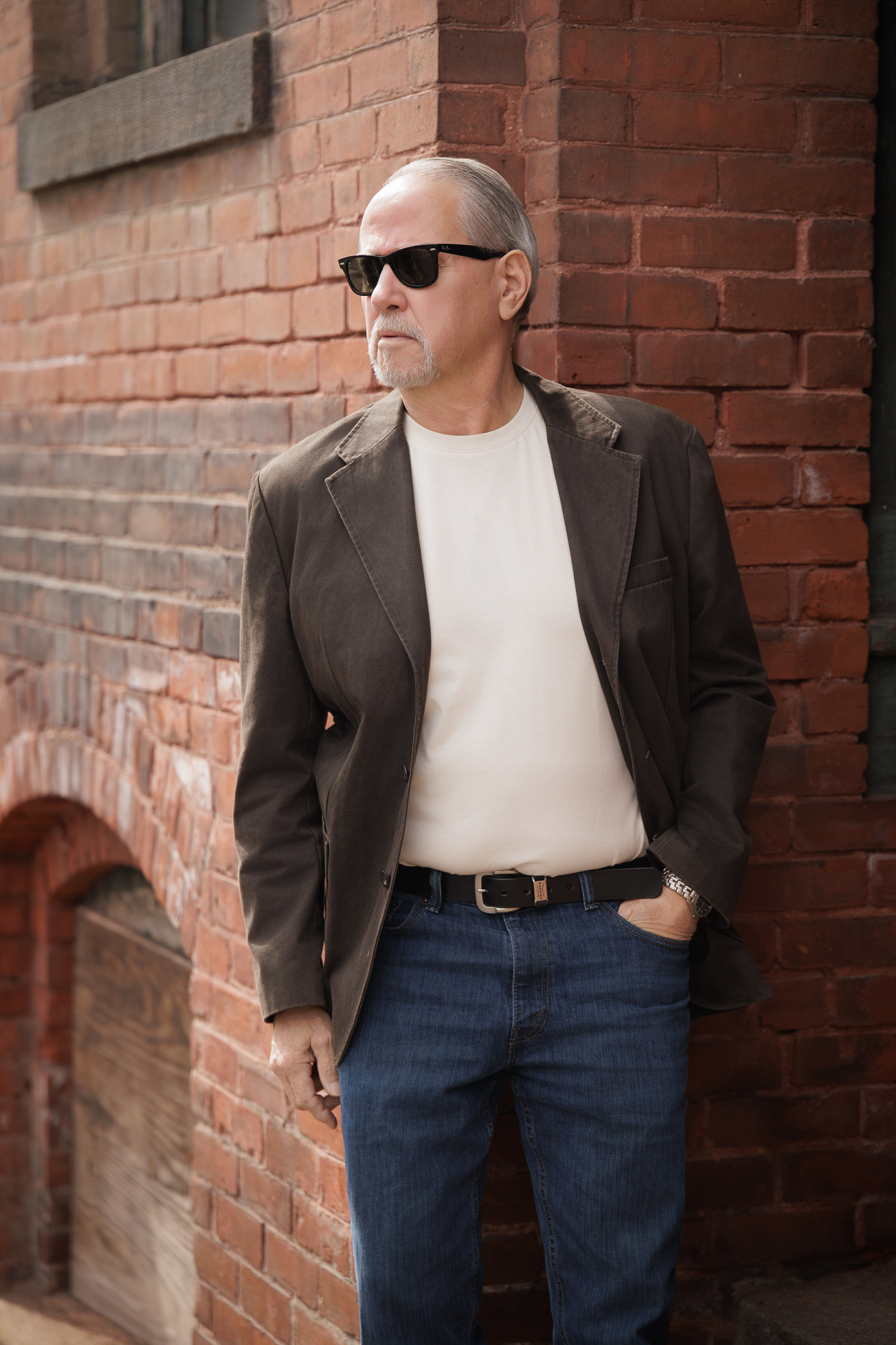 A man seated on stone steps in a white Essential Crew under a brown jacket with jeans