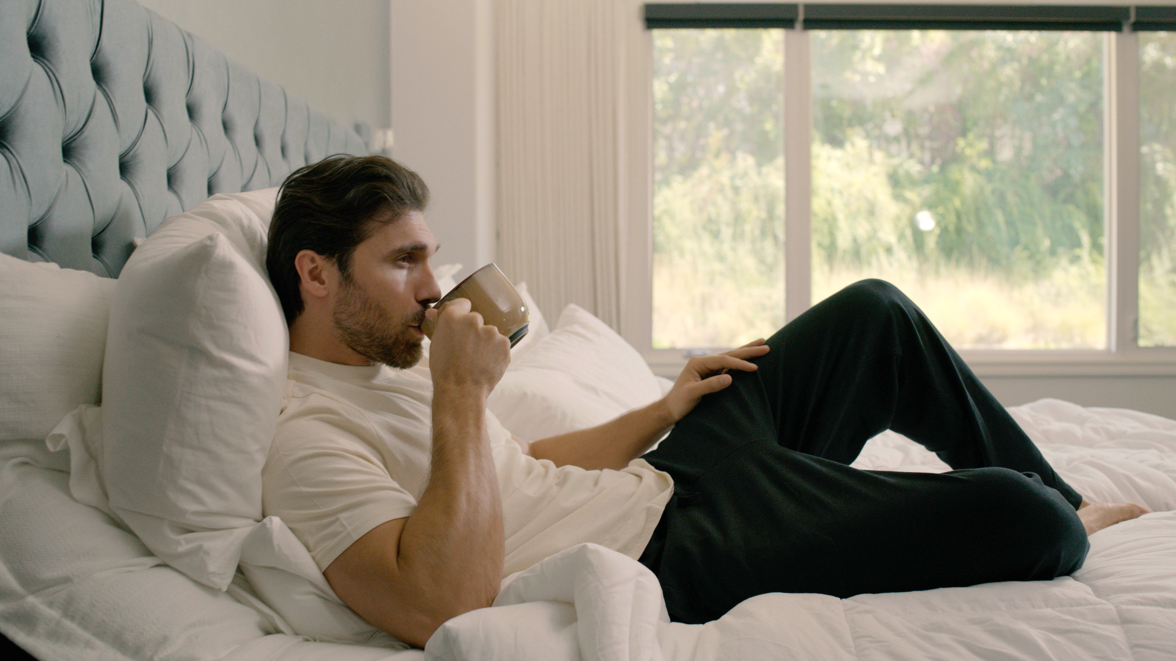 man lounging on a bed drinking coffee dressed in Egyptian Cotton Tshirts' clothing; he is very comfortable