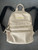 Steve Madden Cream Logo Backpack front flay lay showing black shoulder straps, tp handle, gold Steven Madden plaque