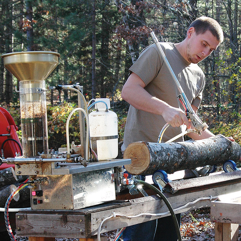Man using the "Pegasus" Semi-Automatic Log Tool in an outdoor setting, inoculating and sealing a mushroom log in one pass