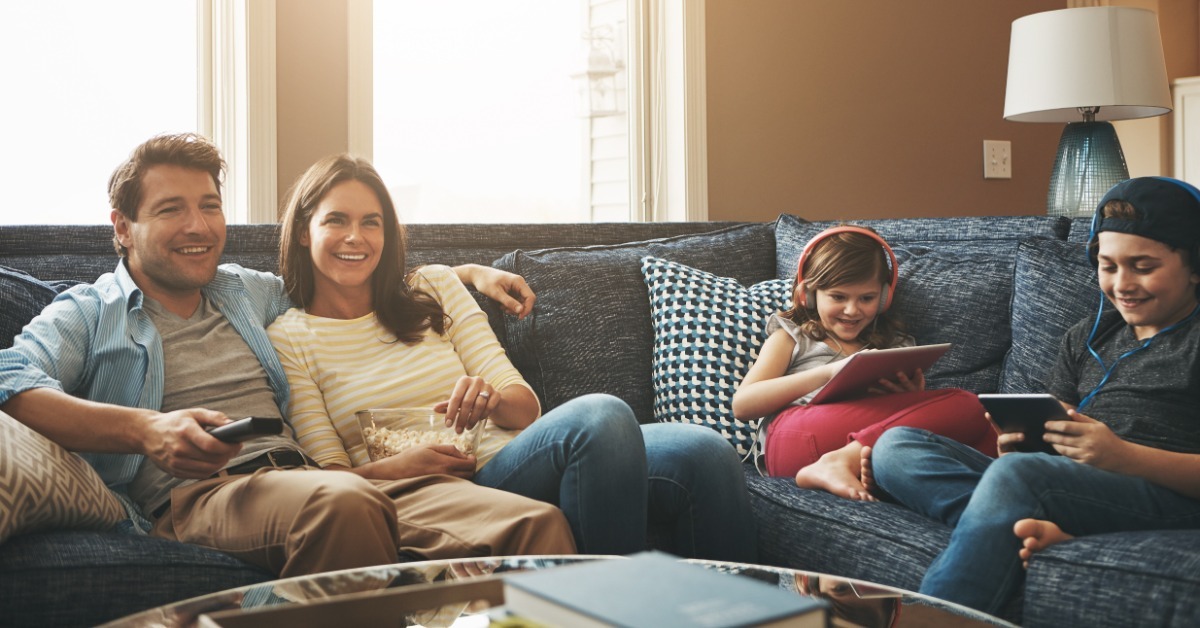 A family sits together on a dark blue sectional sofa. The parents watch TV while the two kids use handheld devices.
