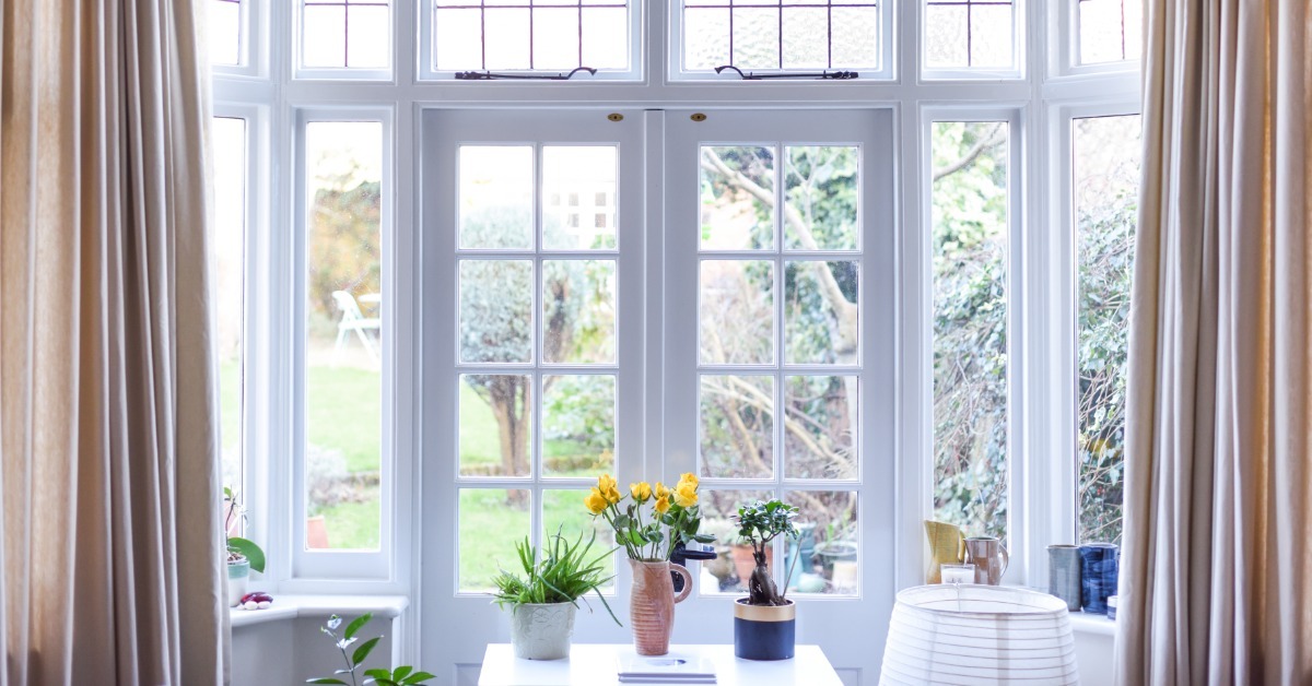 Tall beige curtains frame a large bay window with white mullions and glass-paneled doors. Plants are on a nearby table.