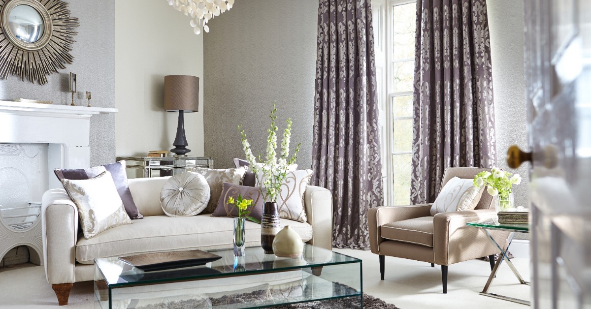 A neutral living room is furnished with a cream sofa, a taupe chair, and patterned purple-gray curtains.