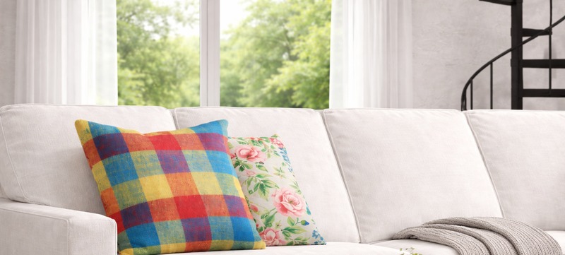 White sectional sofa with colorful plaid and floral pillows in a bright room with sheer-curtained window, soft green outdoor view, and spiral staircase, representing P/Kaufmann decorative fabrics.