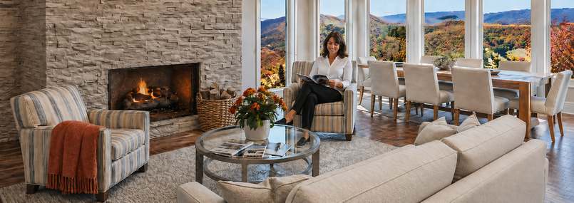 Sunbrella indoor and outdoor performance upholstery fabrics shown in a neutral living and dining room with a linen-look sofa, striped accent chairs, upholstered dining chairs, stone fireplace, and a fall mountain view through large windows.