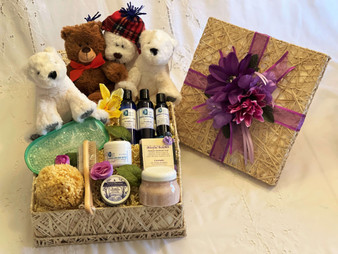 Square wicker basket filled with bath and body products, including blue bottles, sea wool sponge, and a wooden massager, pink Himalayan bath salts. A small teddy bears are leaning against the basket. 