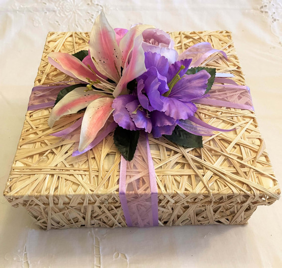 Handmade beige colored Lupis basket with Lid decorated with silk flowers and ribbons in pink, white and purples hues. Approximate size 10 x 10 x 3.5 inches.