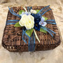 Dark brown basket decorated with blue and white silk flowers, green leaves and blue and white ribbon. 