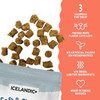 A product infographic for Icelandic+ cat treats. The left side features a white background showing small, square brown treat pieces spilling upward out of an open bag that displays the top gray and white herringbone pattern banner and the "ICELANDIC+" brand label. The right side has a coral-colored panel with a wavy border containing a vertical list of product features in white text, each accompanied by a simple line icon. The listed features are: "3 CALORIES PER TREAT" with the number 3, "PACKED WITH FLAVOR CATS LOVE" with a cat face icon, "NO ARTIFICIAL FILLERS OR PRESERVATIVES" with a plant sprout icon, "AIR DRIED, LIMITED INGREDIENTS" with a heat wave icon, and "MADE IN SMALL BATCHES" with a heart icon.