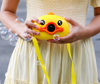 A close-up lifestyle photograph of a child holding a yellow duck-shaped bubble machine. The child is wearing a light yellow smocked dress and holds the camera-style toy with both hands at waist level. The toy features large black cartoon eyes, an orange duck beak surrounding the central bubble nozzle, and a bright yellow carrying strap hanging down. Several clear, iridescent soap bubbles float in the air around the child's hands and the toy against a softly blurred grey background.