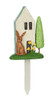A front product photograph of a single miniature decorative garden house pick isolated against a white background. The resin stake features a tall off-white cottage with a light teal roof trim, a large brown rabbit on the left, yellow flowers by the door, and a slender green tree on the right, all resting on a green grass base with a white ground stake.