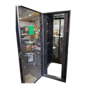 A front view of an open black APC server rack enclosure with its perforated mesh front door swung completely open, revealing a hollow internal frame and a warehouse background, isolated on a black background