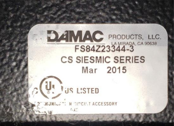 A close-up photograph of a silver metal identification plate on a textured black surface, showing the DAMAC Products logo, model number FS84Z23344-3