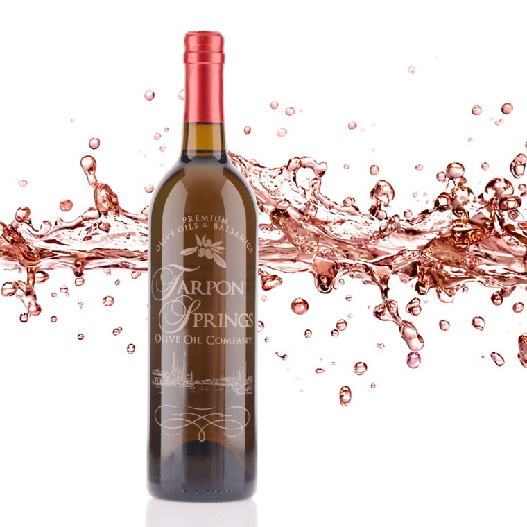 Tarpon Springs Olive Oil Company Rosé Balsamic in a tall dark glass bottle with red foil capsule, etched brand logo and harbor scene, surrounded by a dynamic splash of rose-colored balsamic on a white background.