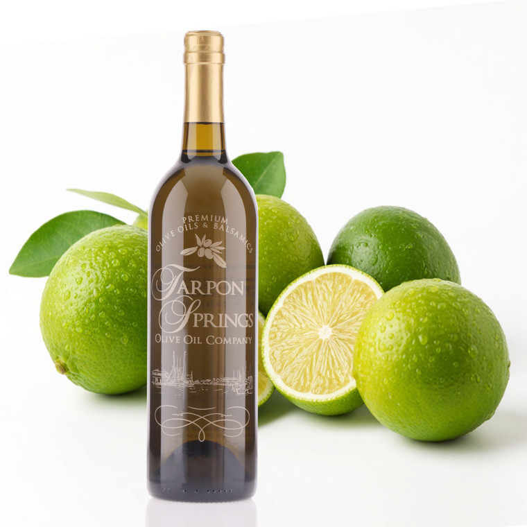 Product photo of Tarpon Springs Olive Oil Company premium green lemon extra virgin olive oil. A 750ml dark glass bottle with gold foil cap and white etched label stands in front, with two larger bottles shown as faint overlays and a small sample bottle on the right. Behind the bottles are fresh green lemons with water droplets, green leaves, and one lemon sliced in half to show the juicy interior, all on a clean white background.