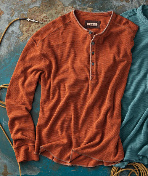 32 Bar Blues Once Around The Park, folded rust-colored long-sleeve shirt with a button-up collar and textured fabric, placed on a rustic surface with a rope and a partially visible teal shirt.