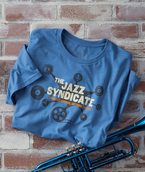 32 Bar Blues All We Need Is A Place T-Shirt, Blue T-shirt with jazz syndicate logo and music-themed graphics, placed on a brick wall background.