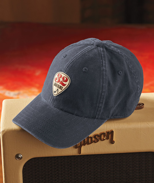 32 Bar Blues Kick In The Head, image of a navy blue baseball cap with the 32 Bar Blues logo on the front, placed on a guitar amplifier with a blurred warm background.