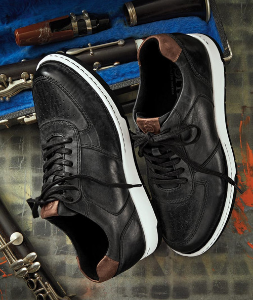 32 Bar Blues Don't Stop Me Now, black leather casual sneakers with brown accents and white soles placed on a surface with a saxophone and musical instrument case in the background.