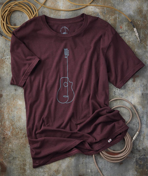 32 Bar Blues Big Six, maroon T-shirt with a minimalist line drawing of a guitar with a comb attached to its neck, laid out on a textured concrete surface along with a coiled gold-colored cable and a pair of vintage-style headphones.