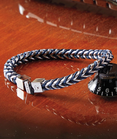 32 Bar Blues Twisting Waves bracelet, close-up of a stylish woven navy and white cord bracelet with metallic accents resting on a wooden surface beside a timer.