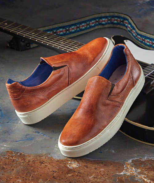 32 Bar Blues Road Dog, stylish brown leather slip-on sneakers with white soles and blue interior, placed on a textured surface with a guitar in the background.