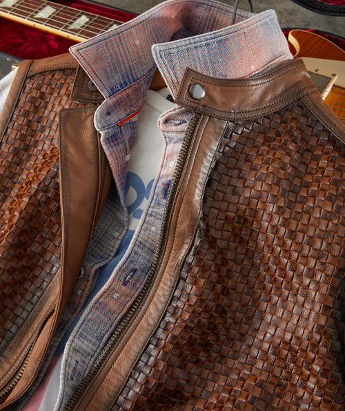 32 Bar Blues Hello Number One, close-up of a brown leather jacket with woven textured pattern, paired with a plaid shirt and a graphic t-shirt, and a guitar in the background.