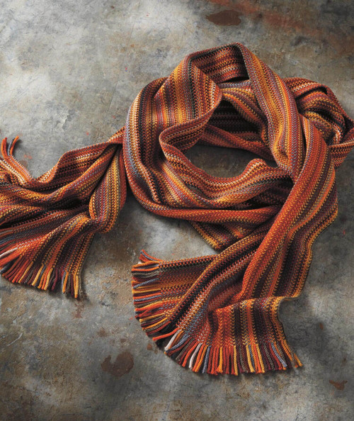 Quiet Fire, a colorful woven scarf with orange, red, black, and brown stripes, draped on a rustic concrete floor.