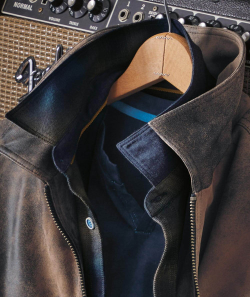 All Out, black leather jacket with a partially open front showing a blue and black striped shirt inside, placed on a hanger with a background of sound equipment and speaker grills.